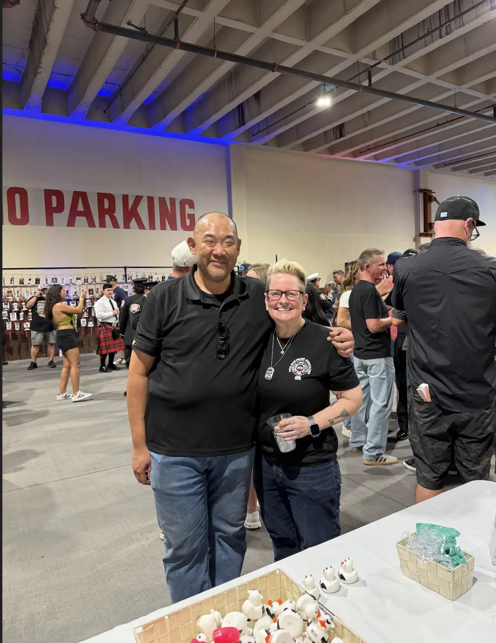 Two smiling attendees at the indoor 9/11 tribute gathering, with a memorial wall of first responder portraits behind them